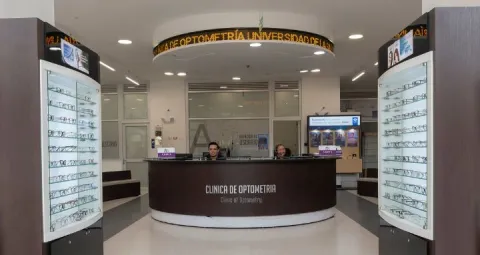 Recepción de la clínica de optometría. Hay dos vitrinas llenas de gafas a ambos lados del mostrador de recepción. Detrás del mostrador, hay dos personas sentadas,