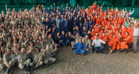 Grupo de personas posando para una foto grupal, llevan uniformes de diferentes colores: beige, azul y naranja. Están de pie y sentados en filas, con algunas personas levantando las manos