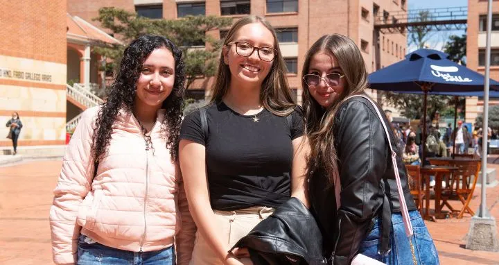 Tres jovenes mujeres de pie al aire libre en un patio con edificios de ladrillo...